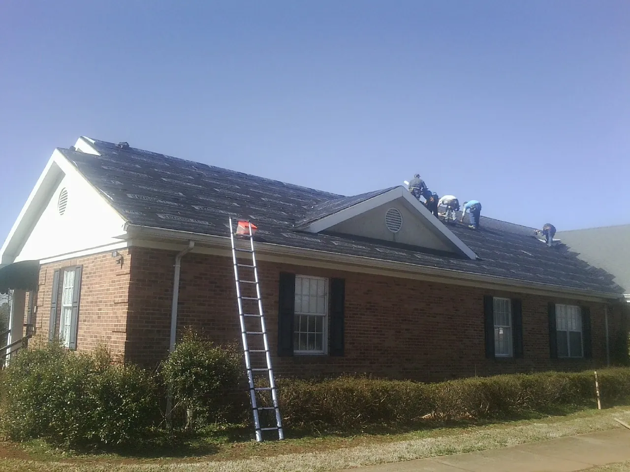 Crew tearing shingles off a brick ranch roof with a ladder set — Emergency Roof Repair work by our crew in Gracemont