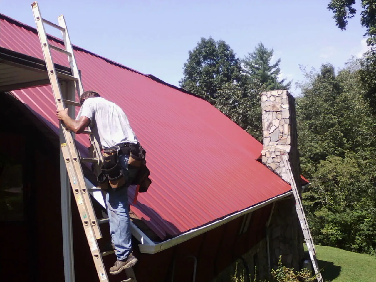 Roofer on a ladder beside a red metal roof and stone chimney — Soffit & Fascia Repair by Roofing Brothers in Gracemont